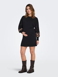 Black knit dress with long sleeves featuring colorful floral embroidery. Model wearing brown knee-high boots, standing in a neutral background.