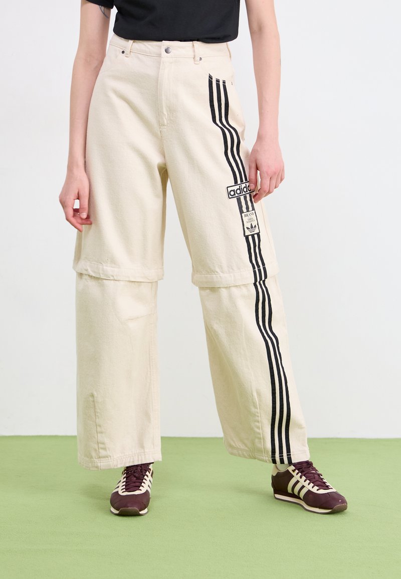 Bež široke hlače s crnim bočnim prugama i Adidas logotipom. Napravljene od čvrstog materijala, s ležernim krojem i skraćenom dužinom.