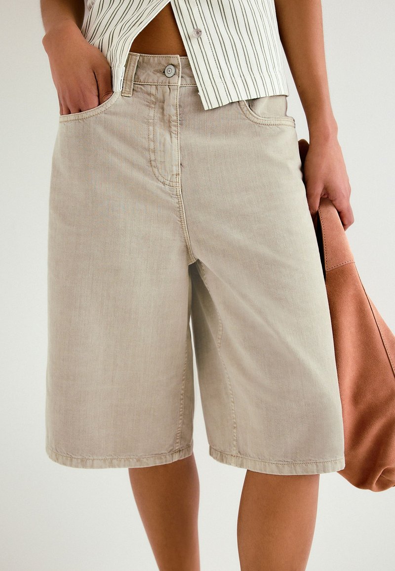Person som bär beige knälånga jeansshorts och en randig kort skjorta, håller en brun mockaväska i ena handen.