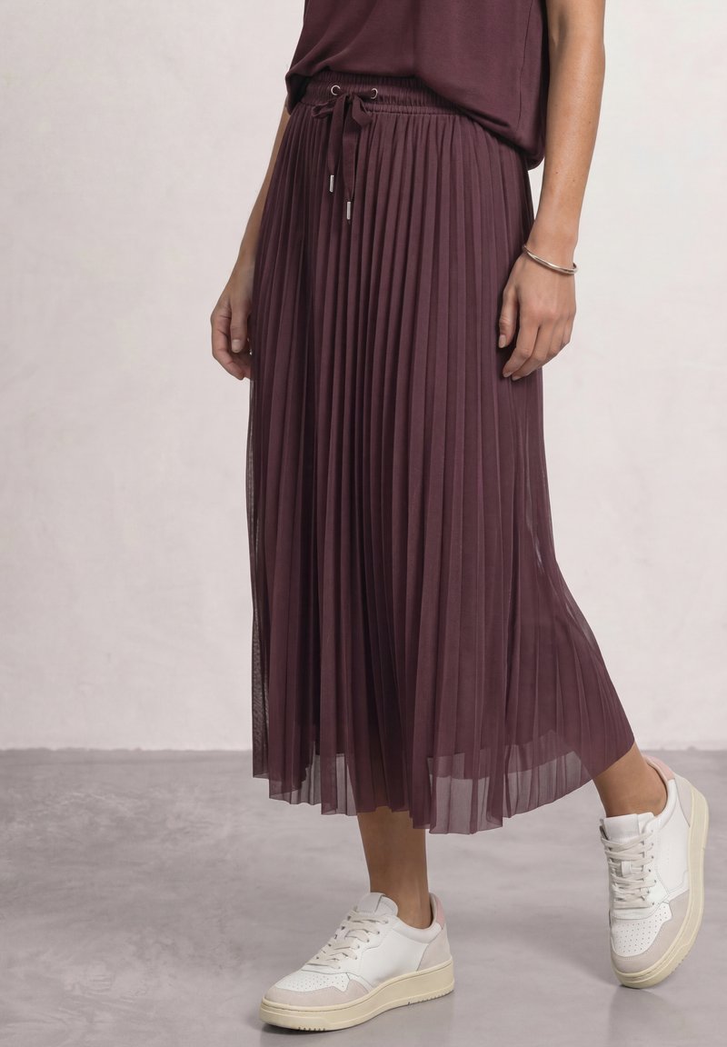 Femme portant une jupe midi plissée bordeaux avec une taille à cordon et des baskets blanches, debout sur un sol gris clair.