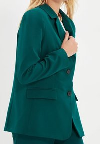 Blazer turchese realizzato in tessuto liscio, caratterizzato da colletto a sfondo, tre bottoni e due tasche laterali, progettato per una silhouette aderente.
