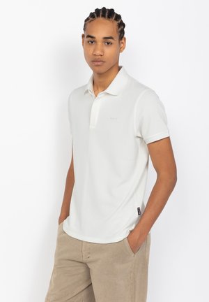 Polo blanc avec une finition texturée, manches courtes et col. Présente un logo discret et une étiquette sur le côté. Associé à un short kaki.