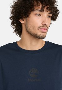 Navyblaue Baumwoll-T-Shirt mit einem Rundhalsausschnitt und einem dezenten, dunklen Logo eines Baumes sowie "Timberland" auf der Vorderseite.