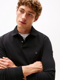 Juodas polo marškinėliai su tekstūruotu audiniu, turi mygtukinę apykaklę ir mažą logotipą ant krūtinės. Modelis pozuoja sukryžiuotomis rankomis.