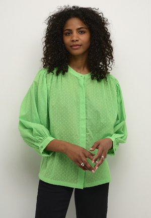 Green, dotted sheer blouse with a button-up front, puffed long sleeves, and gathered shoulders, paired with black pants and rings.