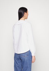 Camisa branca de botões com gola plissada, mangas longas e formato ajustado, acompanhada de jeans de ganga azuis com bolsos traseiros.