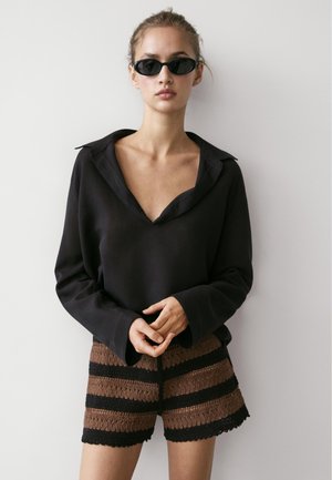 Kobieta ubrana w czarną bluzkę z głębokim dekoltem w serek i długimi rękawami, brązowe i czarne paskowane dzianinowe szorty oraz czarne owalne okulary przeciwsłoneczne, stojąca na tle białej ściany.