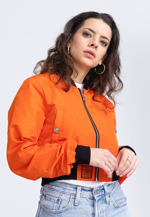 Pomarańczowa kurtka bomber z czarnymi mankietami i dołem, z zamkiem błyskawicznym oraz kieszenią z przodu. Cechuje się metką na talii. Noszona na jasnym dżinsie.