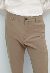 Beige Hosen mit einer glatten Textur, ausgestattet mit einem Reißverschluss, Knopfverschluss und seitlichen Taschen. Dezente Nähte sind sichtbar.