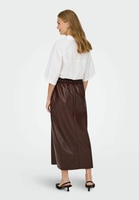 Jupe en faux cuir marron avec une taille élastique, associée à une chemise blanche à manches courtes. Talons noirs à bout ouvert. Texture lisse, longueur genoux.