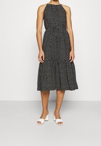 Svart ärmlös klänning med vita prickar, rynkad halsringning, åtdragen midja, flödande kjol, tillsammans med vita klackskor med blockhäl.