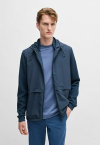 Leichte blaue Zip-Jacke mit Kapuze, strukturierte Ärmel, zwei Fronttaschen, über einem blauen T-Shirt getragen, kombiniert mit blauen Hosen.