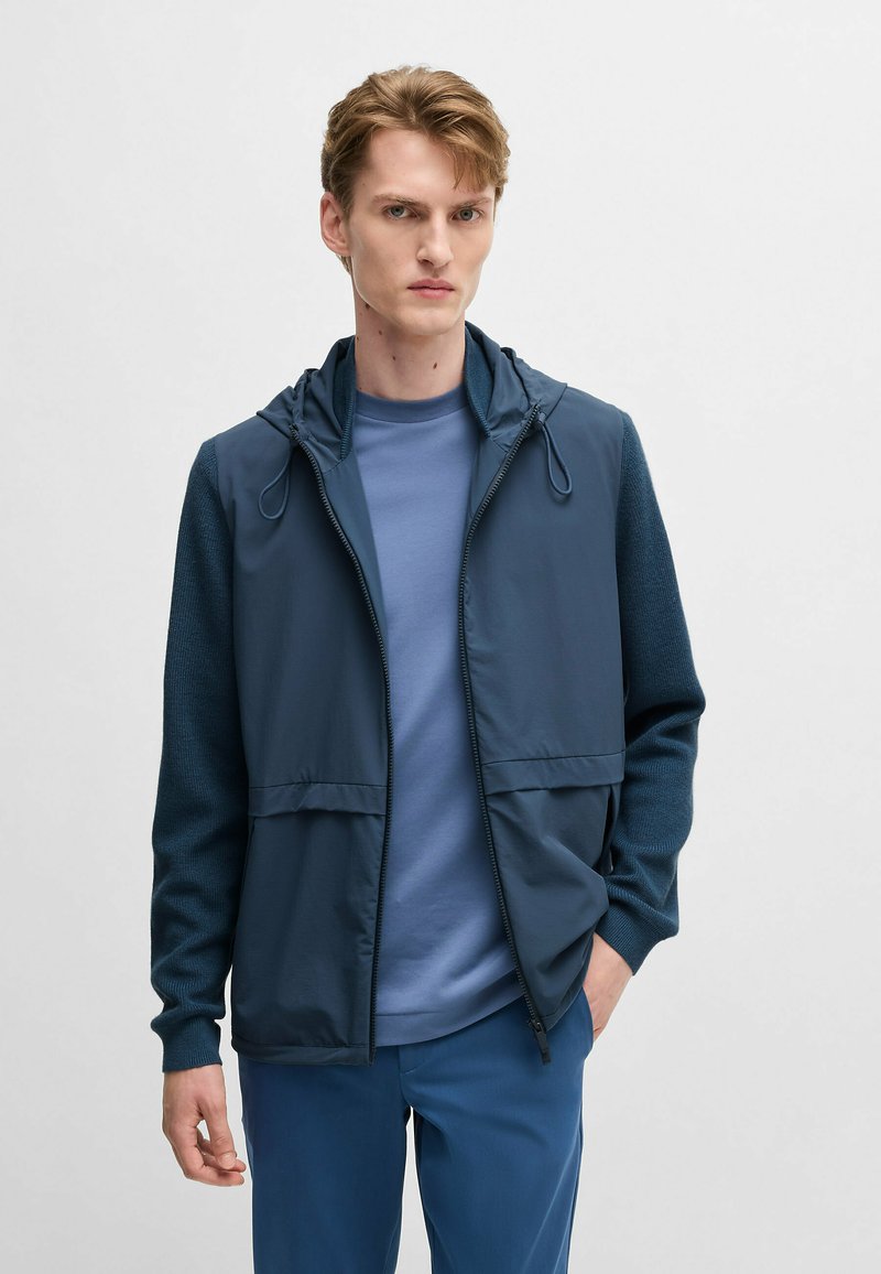 Leichte blaue Zip-Jacke mit Kapuze, strukturierte Ärmel, zwei Fronttaschen, über einem blauen T-Shirt getragen, kombiniert mit blauen Hosen.