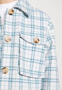 Biała i niebieska kraciasta kurtka z teksturowanej tkaniny, z dużymi brązowymi guzikami i kieszenią na piersi.