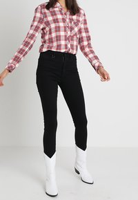 Camicia a quadri rossa e bianca con bottoni e maniche lunghe, abbinata a jeans skinny neri e stivali alla caviglia bianchi.