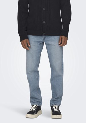 Man draagt lichtblauwe jeans met rechte pijpen, een zwarte geribde gebreide cardigan met knopen en zwarte sneakers met witte zolen en veters.