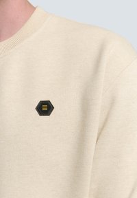 Beige sweatshirt met een textuurstof, voorzien van een zwarte hexagonale patch met gele lijnen op de linkerkant van de borst. Ribbelkraag.