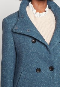 Dvouřadový modrý vlněný kabát s texturovaným povrchem, který má černé knoflíky a klopový límec přes krémový pletený svetr.