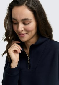 Sweat-shirt zippé bleu marine avec un col haut, texture lisse et couture décorative. Le modèle touche délicatement la fermeture éclair.