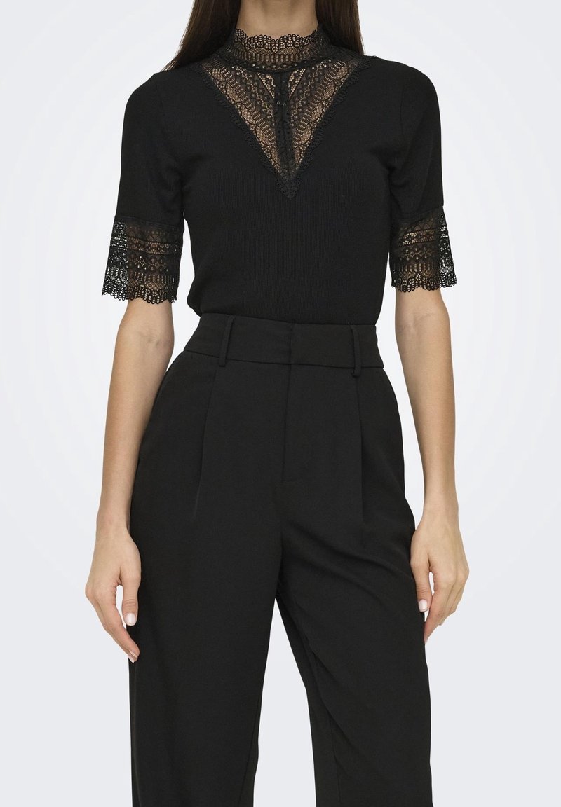 Femme portant un pantalon noir taille haute et un haut noir à manches courtes avec des détails en dentelle sur les manches, le décolleté et le panneau poitrine.