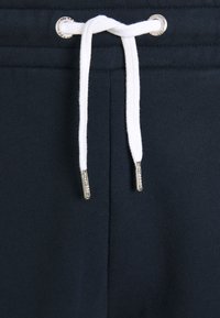 Marineblaue Sweatpants mit einem Kordelzug aus weißen Schnüren und silbernen Metallakzenten. Die Textur ist weich und glatt.