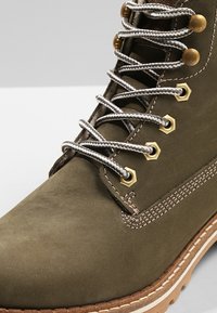 Botte en cuir vert olive avec œillets métalliques, lacets gris et semelle en caoutchouc beige. Présente des coutures contrastées et un design robuste.