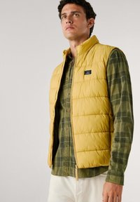 Gilet trapuntato giallo con colletto alto, indossato sopra una camicia a quadri verde. Presenta una chiusura con zip e una toppa con logo sul petto.