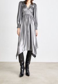 Une robe metallique argentée avec de longues manches bouffantes, un décolleté en V, un buste froncé et un ourlet asymétrique, associée à des bottes noires jusqu'au genou.