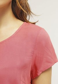 Short-sleeve, scoop-neck top in soft, lightweight fabric, featuring a smooth texture and a solid pink color with subtle shine.
