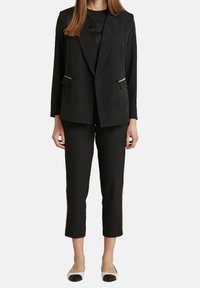 Blazer doppiopetto nero su misura con rever a scomparsa, abbinato a pantaloni corti coordinati con accenti laterali. Indossato con scarpe piatte bianche e nere.