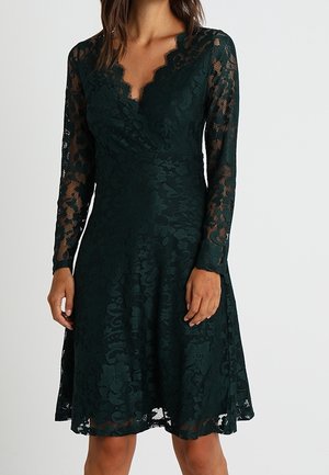 Femme portant une robe en dentelle vert foncé à manches longues avec un décolleté en V festonné, debout devant un fond clair uni.