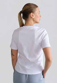 Biały bawełniany t-shirt z krótkimi rękawami, okrągłym dekoltem i prostym dołem, noszony z jasnoszarymi spodniami, widoczny od tyłu.