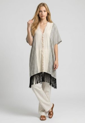 Mujer con cabello rubio largo que lleva un poncho a rayas beige y gris con flecos, pantalones blancos y sandalias marrones, de pie frente a un fondo liso.