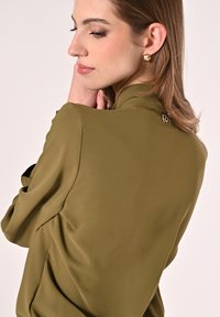 Olivgrüne Bluse mit glatter, glänzender Textur; hat einen hohen Ausschnitt und dezente Raffung, verziert mit einem kleinen metallischen Akzent am Rücken.