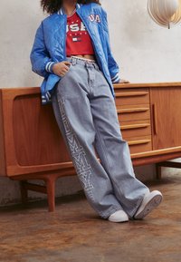 Modrá prošívaná bomber bunda s bílými pruhy, červený zkrácený top, světle modré široké džíny s nápisem "TOMMY" na nohavici a bílé tenisky.