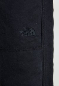 Svart tygplagg med texturerad yta, som har en präglad logotyp "THE NORTH FACE" i det nedre hörnet. Sömdetaljer synliga.