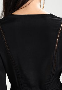 Svart plagg med långa ärmar, med genomskinliga mesh-detaljer längs bakseamens sömmar och en slät, något glänsande tygtextur.