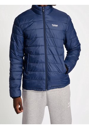 Homme portant une veste matelassée bleu marine zippée et un pantalon de survêtement gris clair, les mains dans les poches de la veste, sur un fond uni.