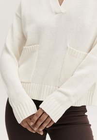 Hvid strikket sweater med dyb V-udskæring, ribbet mansjetter og to forlommer med et struktureret mønster. Bæret over mørke, tætsiddende bukser.