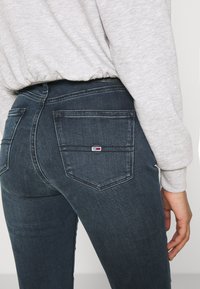 Närbild på person som bär mörkblå jeans i denim och en ljusgrå sweatshirt, med bakficka som visar en liten rektangulär logotypmärke.