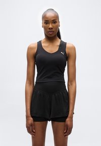 Γυναίκα με μαύρο αθλητικό τοπ και σορτς, στέκεται με το πρόσωπο προς τα εμπρός σε απλό λευκό φόντο.