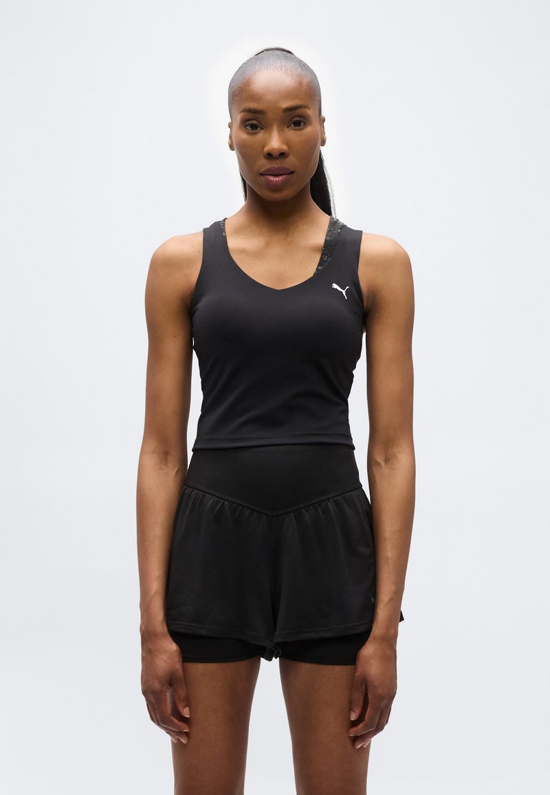 Γυναίκα με μαύρο αθλητικό τοπ και σορτς, στέκεται με το πρόσωπο προς τα εμπρός σε απλό λευκό φόντο.