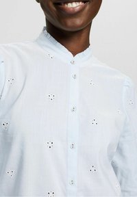 Camicia azzurro chiaro con colletto arricciato, caratterizzata da motivi floreali ricamati e maniche corte. Texture del tessuto morbida.