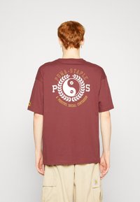Bordo kokvilnas T-krekls ar lielu baltu jinstmaņu grafiku un "PUMA STAPLE" tekstu zeltā uz muguras, īsām piedurknēm, brīvā griezuma.