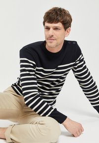 Maglione a righe blu navy e crema in maglia, con scollatura tonda e dettagli con bottoni sulla spalla, abbinato a pantaloni beige chiaro.