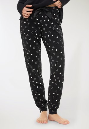 Persona che indossa pantaloni da pigiama neri con motivi a cuori bianchi, cintura in vita legata e scalza su sfondo bianco.