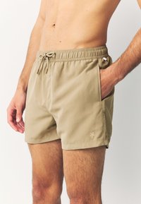 Shorts de bain beiges avec une taille élastique, un cordon de serrage ajustable, des poches latérales et un logo discret en forme de palmier. Texture de tissu lisse.