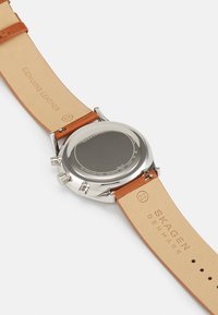 Orologio in acciaio inossidabile argentato con cinturino in vera pelle marrone. Caratteristiche includono una cassa rotonda e tre pulsanti laterali. Nome del marchio inciso.