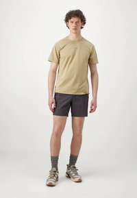 T-shirt bege de algodão e calções escuros, meias cinzentas até ao tornozelo, e sapatos de trilha bege com detalhes roxos, em pé sobre um fundo simples.