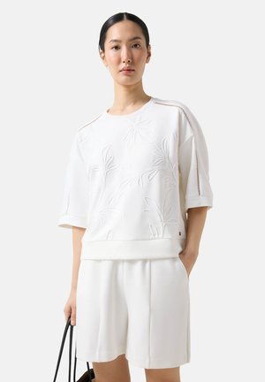 Kobieta ubrana w białą bluzkę z krótkim rękawem o kwiatowym wzorze i dopasowane białe szorty, trzymająca czarną torebkę na pasku, stoi na jednolitym tle.